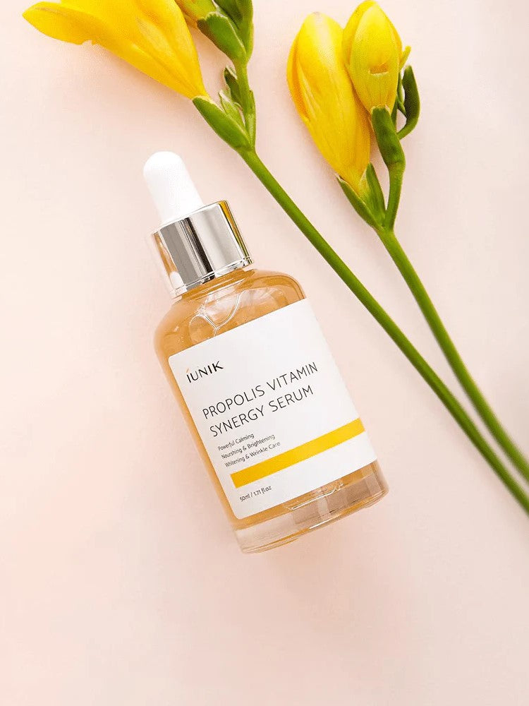Bottle of iUNIK Propolis Vitamin Synergy Serum on a soft pink background with yellow flowers and green leaves, highlighting its natural and nourishing formula.