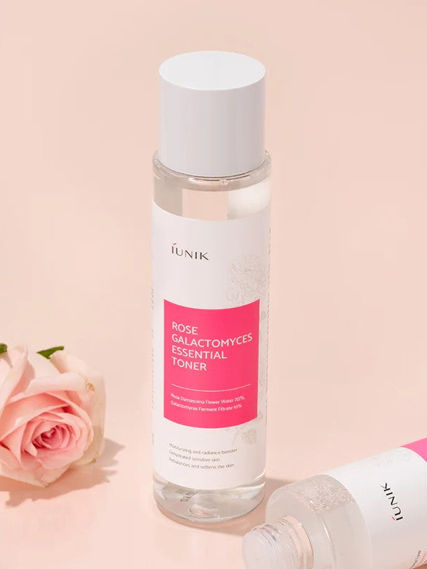 Bottle of iUNIK Rose Galactomyces Essential Toner with pink label next to a delicate pink rose on a soft pastel background, emphasizing gentle hydration effects.