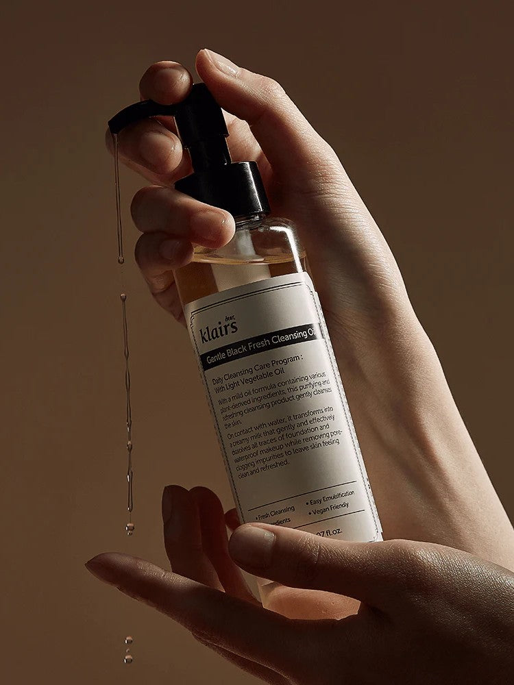 A hand holding Klairs Gentle Black Fresh Cleansing Oil with droplets falling, set against a warm brown background, emphasizing its lightweight texture.