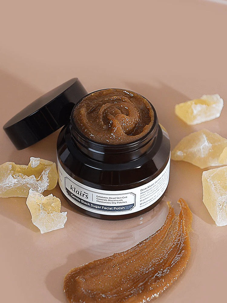 Klairs Gentle Black Sugar Facial Polish jar open to display its rich brown exfoliating scrub, surrounded by sugar crystals on a smooth, light surface, emphasizing its gentle exfoliation.