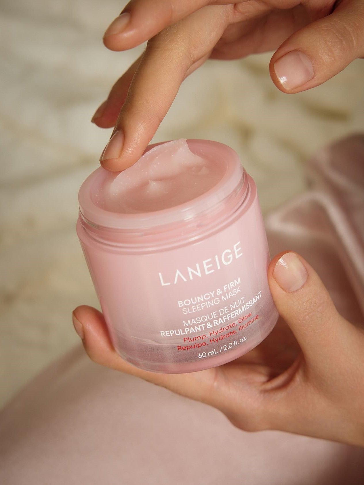 Close-up of a hand holding the Laneige Bouncy & Firm Sleeping Mask pink jar, while two fingers of the other hand dip into its creamy texture.