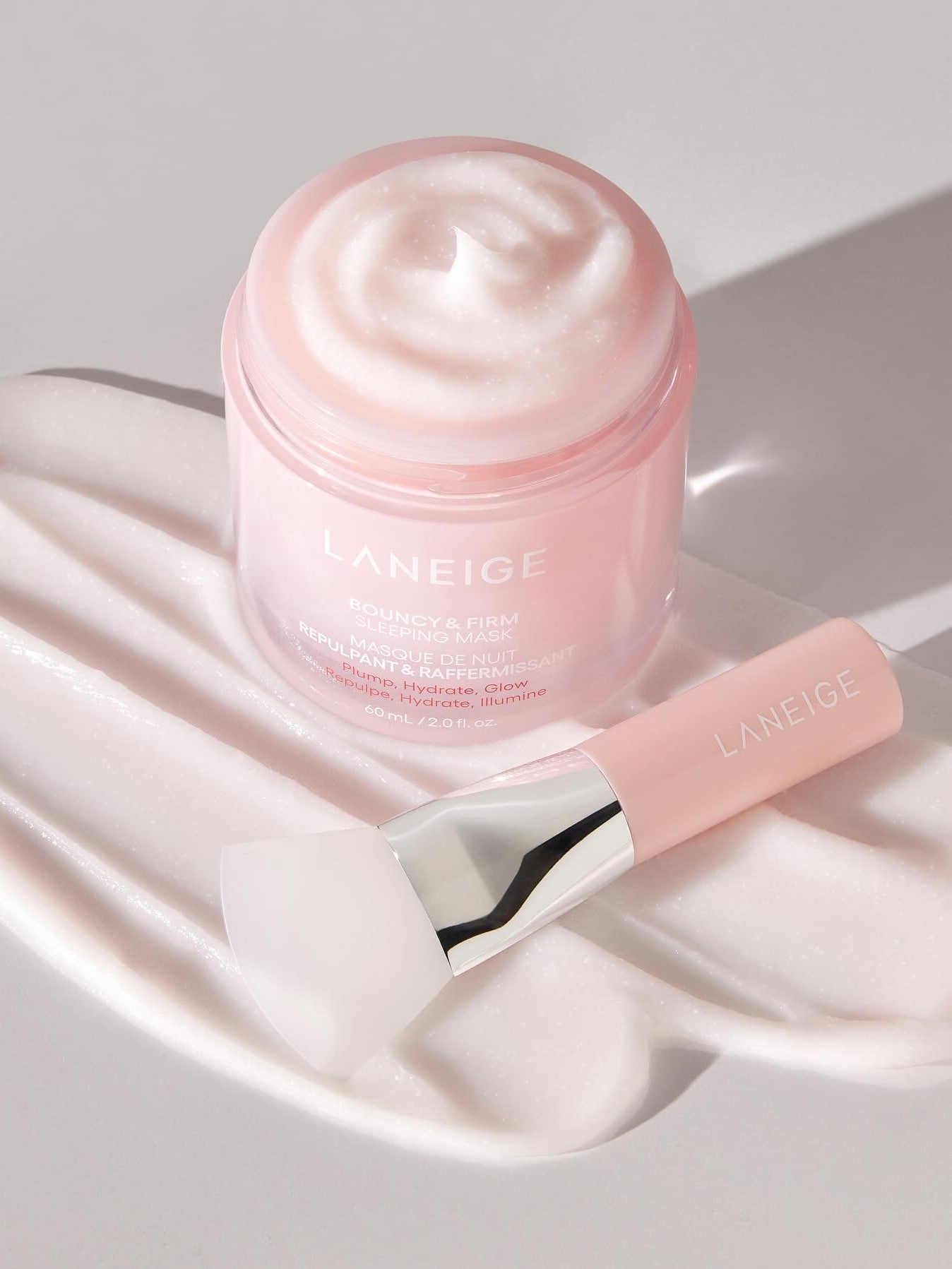 Open pink jar of Laneige Bouncy & Firm Sleeping Mask with creamy texture and matching applicator on a soft pink background.