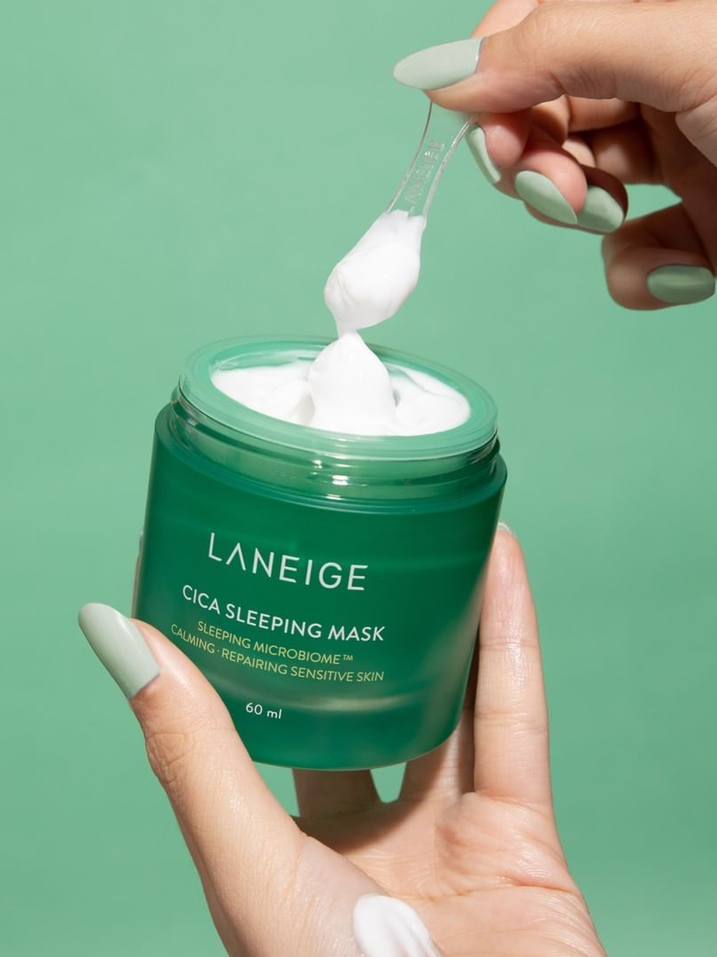 Two hands holding a green jar of Laneige Cica Sleeping Mask, with a plastic spatula scooping creamy white texture against a matching green background.