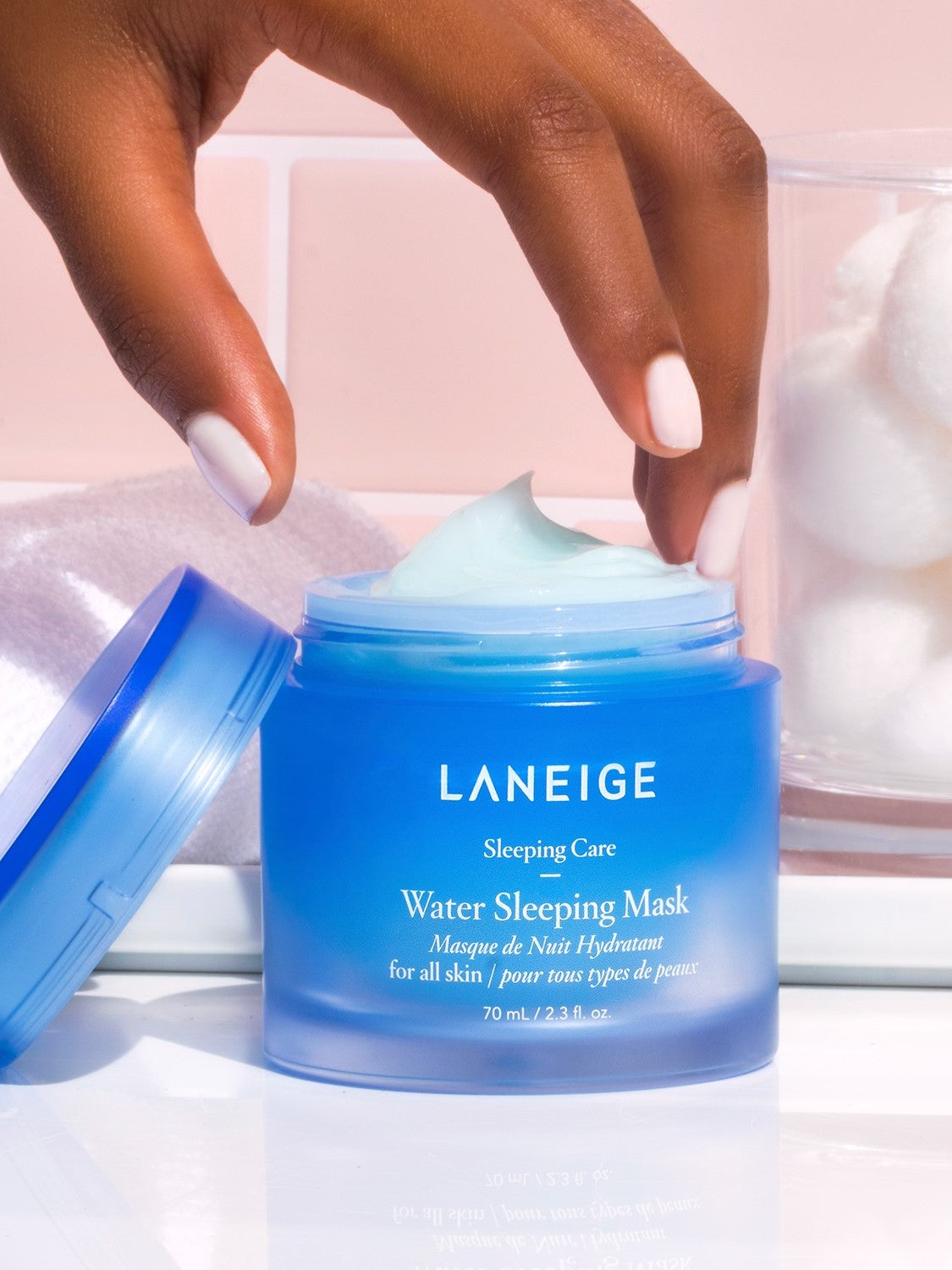 A hand reaching for the open blue jar of Laneige Water Sleeping Mask, with cotton balls and a soft towel in the background, highlighting its lightweight gel texture.