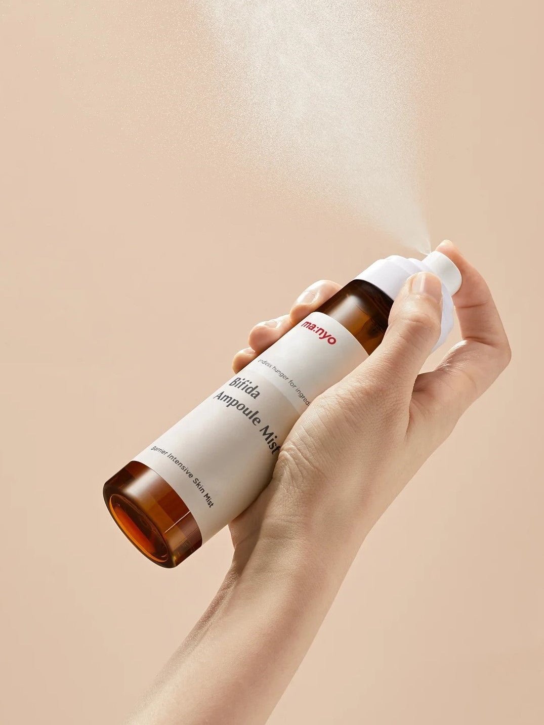 Hand holding a bottle of Manyo Bifida Ampoule Mist releasing a fine mist against a soft beige background, highlighting its lightweight and hydrating formula.