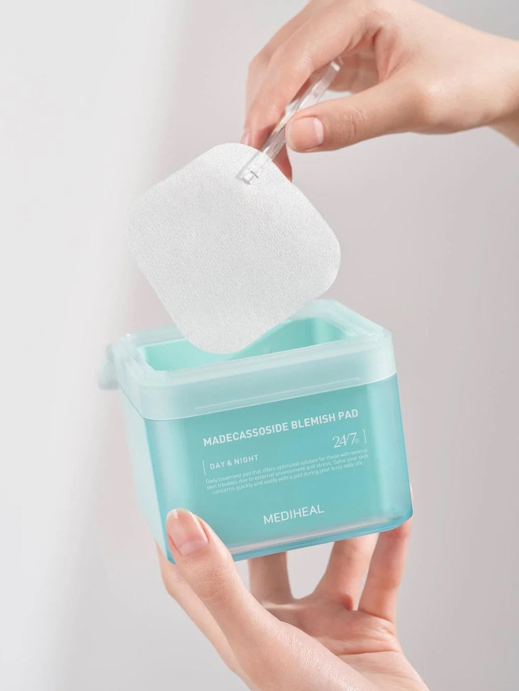 A hand holding a turquoise container of Mediheal Madecassoside Blemish Pad, lifting a cotton pad out with a clear tool, showcasing ease of use.
