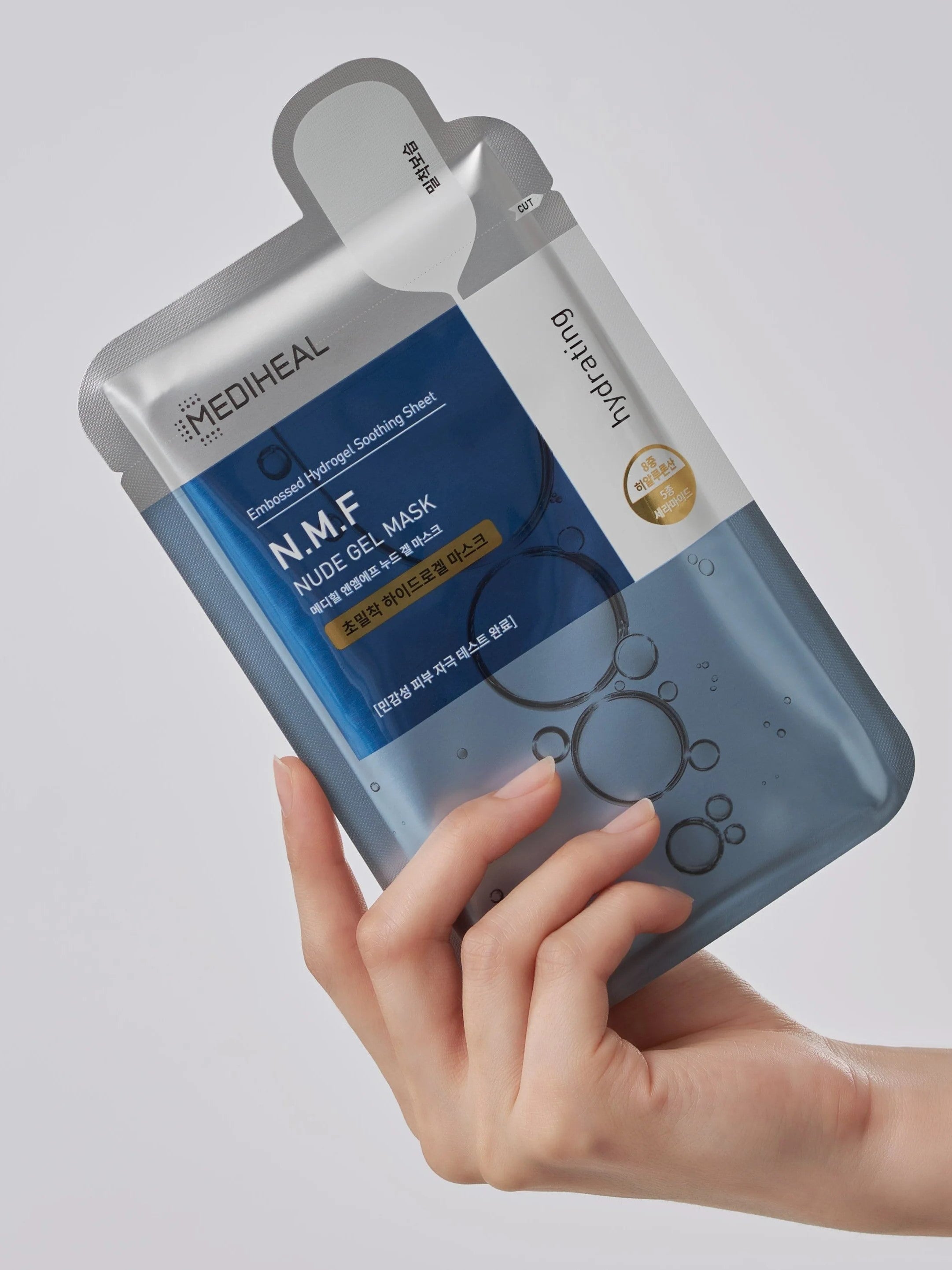 A person holds a Mediheal N.M.F Nude Gel Mask packaging with a blue design, emphasizing hydration and soothing benefits.