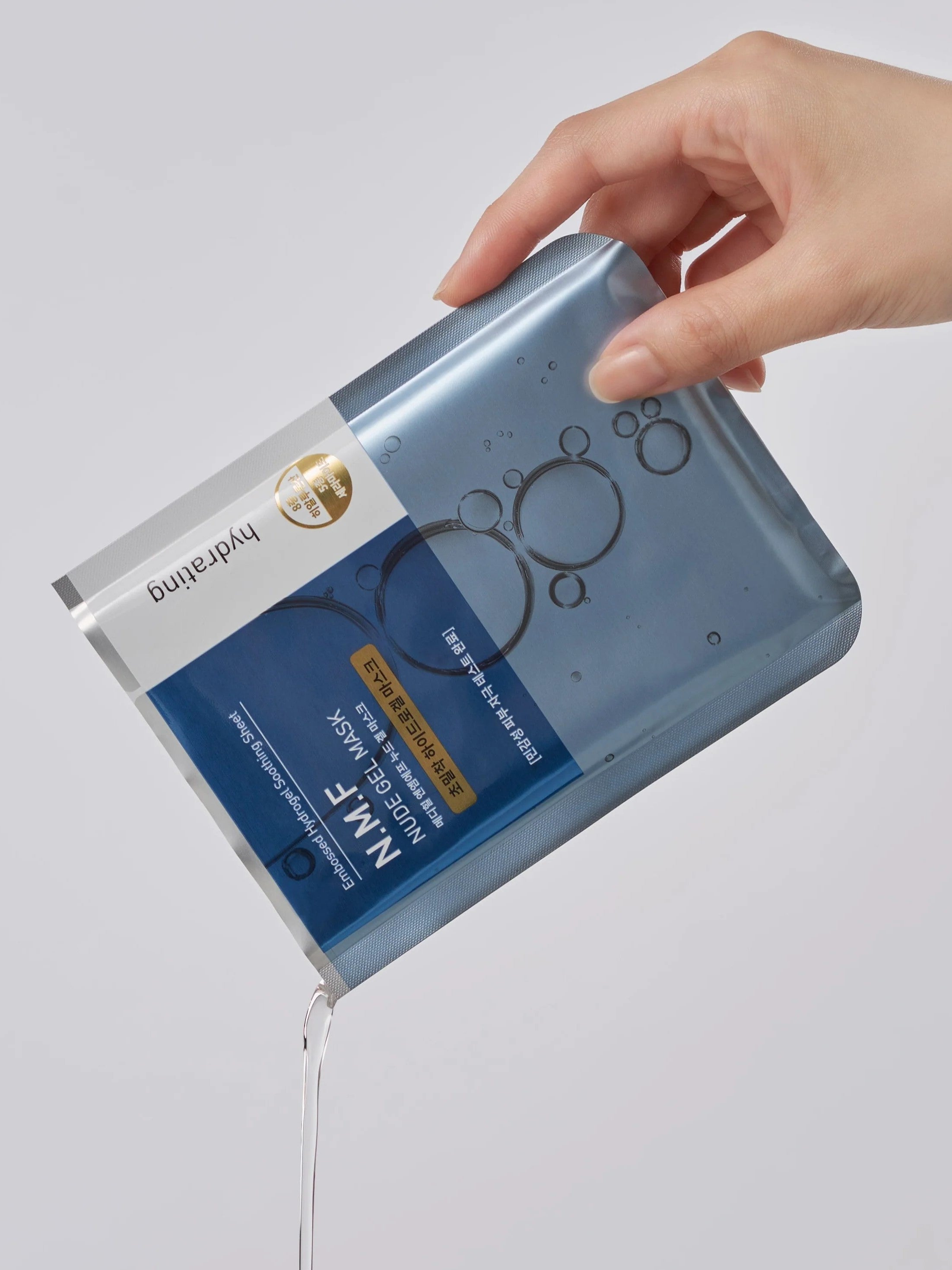 A hand holds the Mediheal N.M.F. Nude Gel Mask package, with essence spilling out to highlight its hydrating benefits.