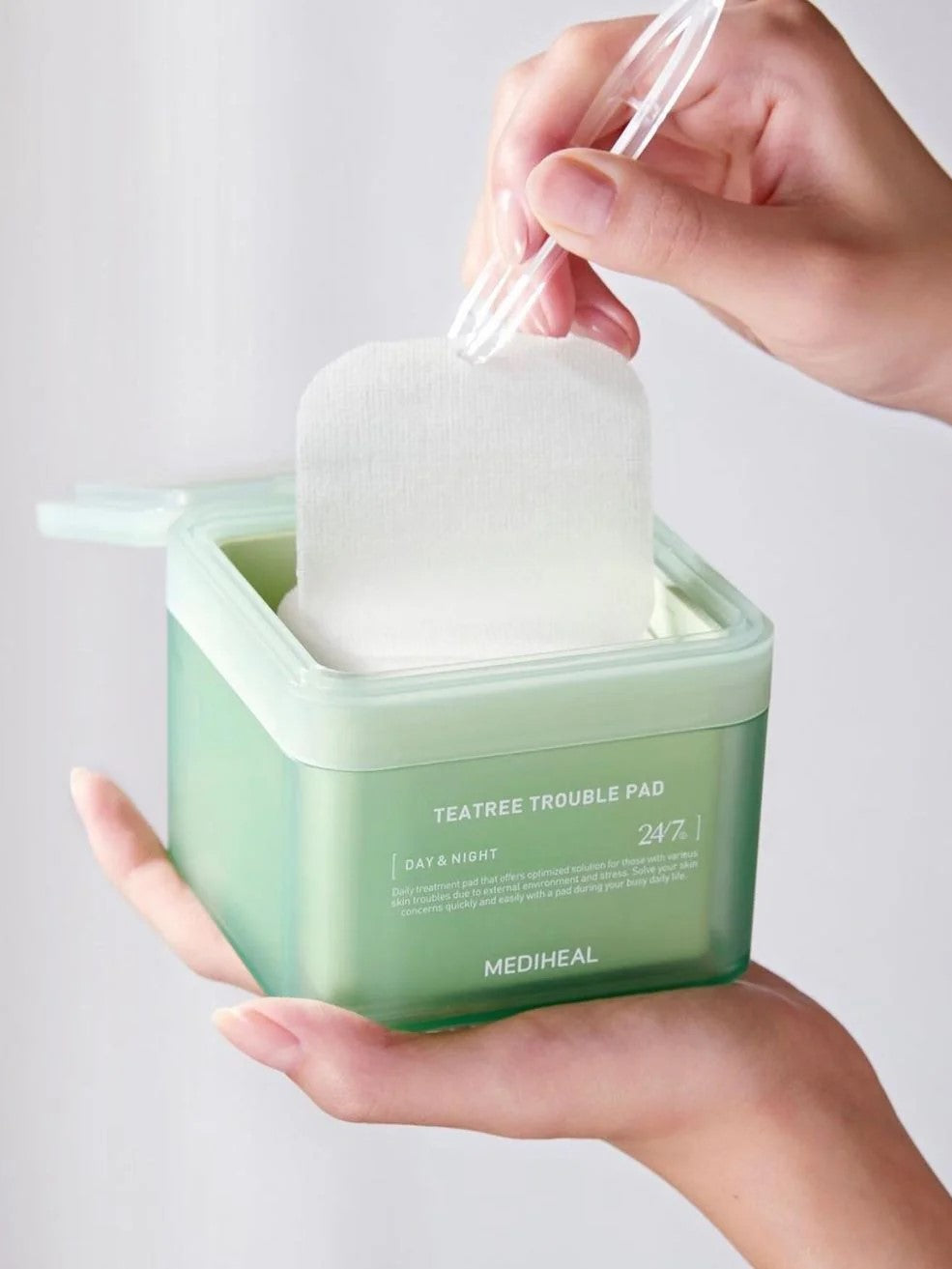 A hand holding a green container of Mediheal Teatree Trouble Pad, lifting a cotton pad out with a clear tool, showcasing ease of use.