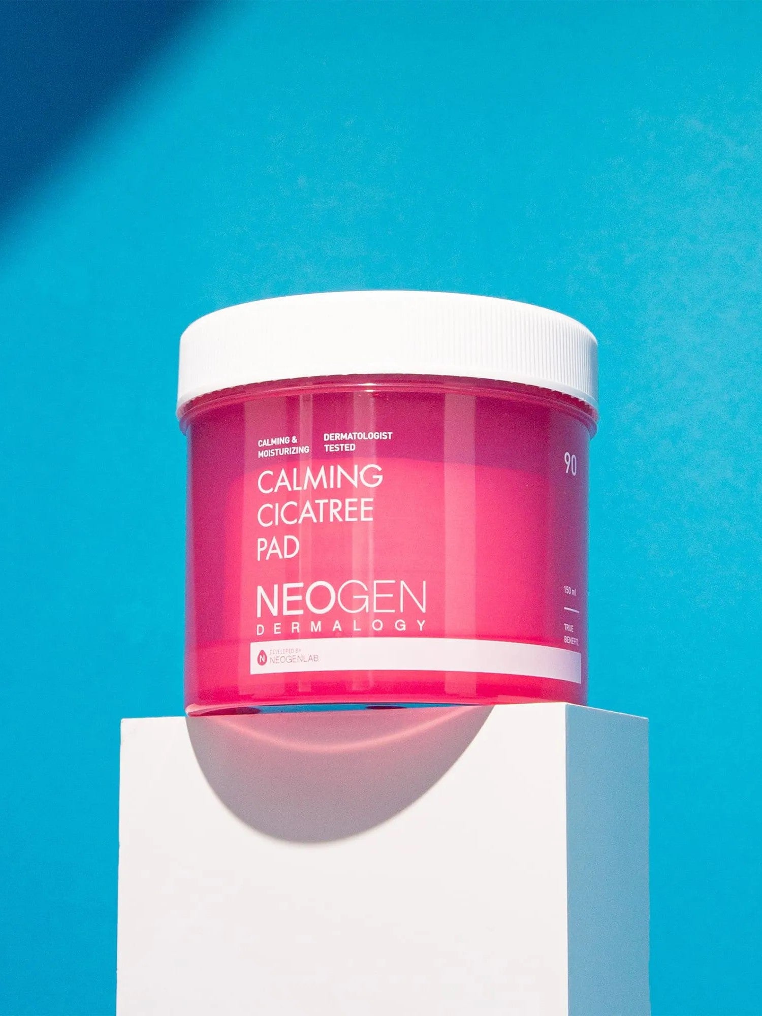 Bright pink jar of Neogen Dermalogy Calming Cicatree Pad displayed on a white pedestal against a vibrant blue background, highlighting its soothing benefits.