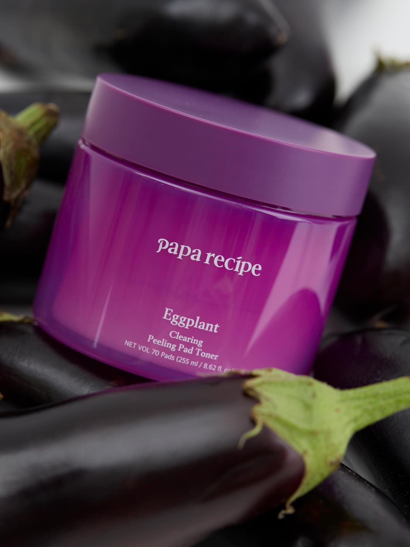 A purple jar labeled 'Papa Recipe Eggplant Clearing Peeling Pad Toner,' surrounded by fresh eggplants on a dark background, emphasizing natural ingredients.