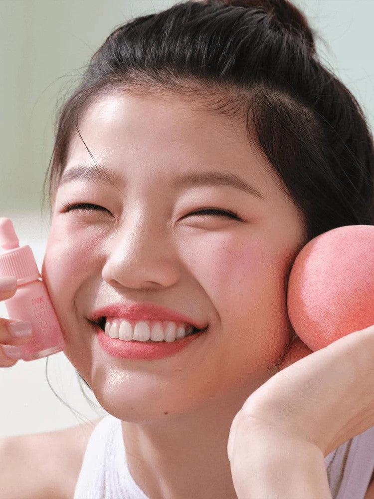 Model holding Peripera INK AIRY VELVET 23 Peaches Collection lip tint in one hand and a peach-colored beauty sponge in the other, against a light background.