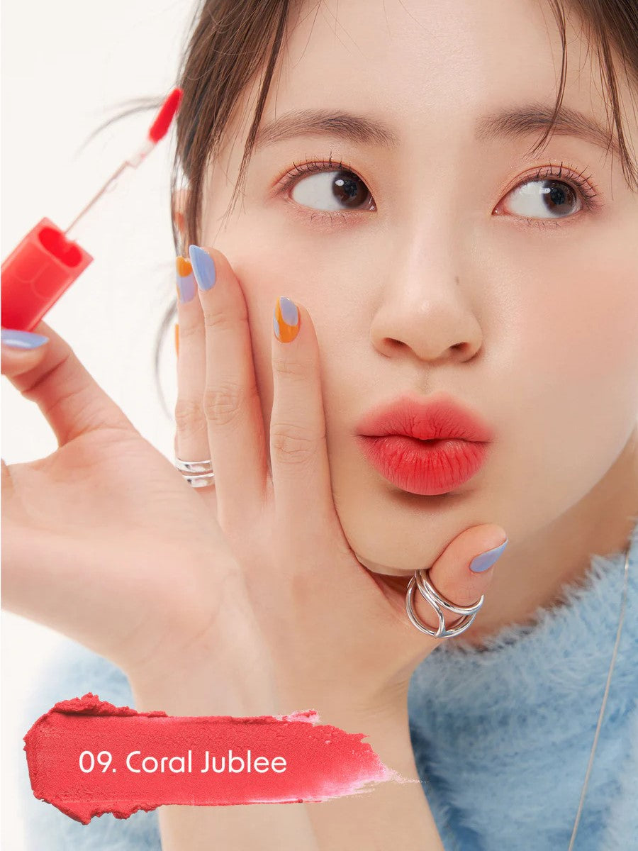 A model wearing Romand Blur Fudge Tint in the shade Coral Jublee, showcasing the soft matte texture on her lips.