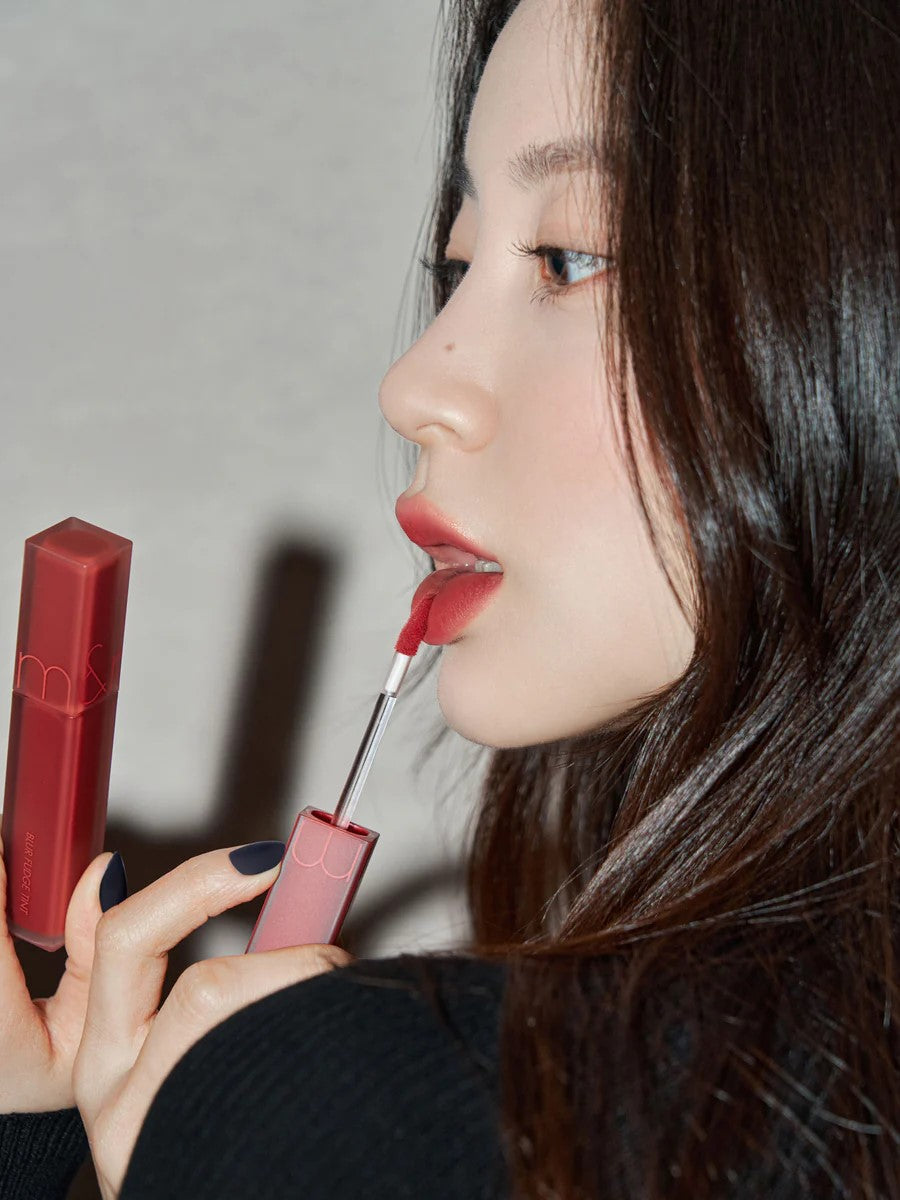 A model wearing Romand Blur Fudge Tint in the shade Currant Jam, showcasing the soft matte texture on her lips.