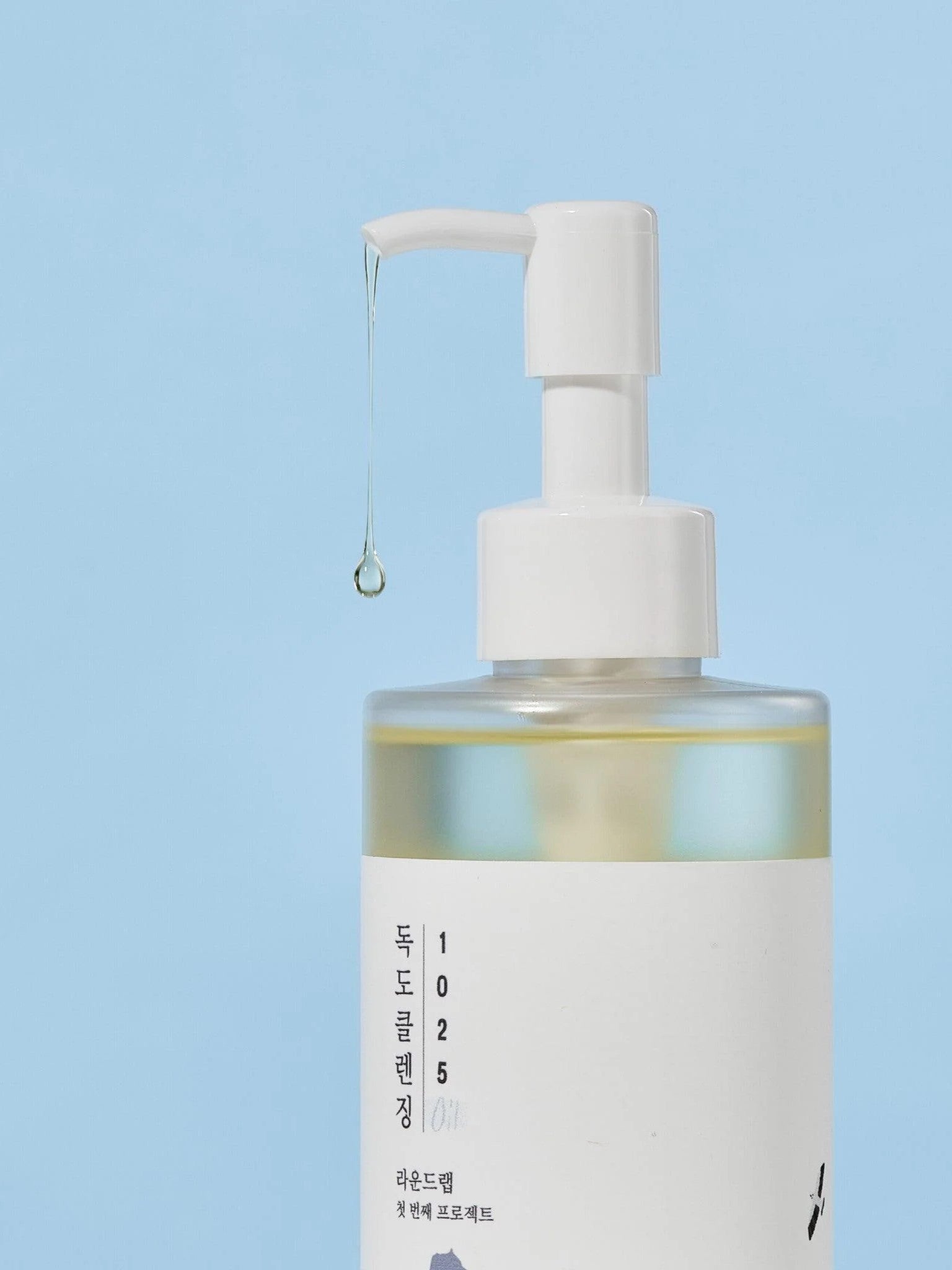 Round Lab 1025 Dokdo Cleansing Oil with liquid dripping from the nozzle, set against a soft blue background.