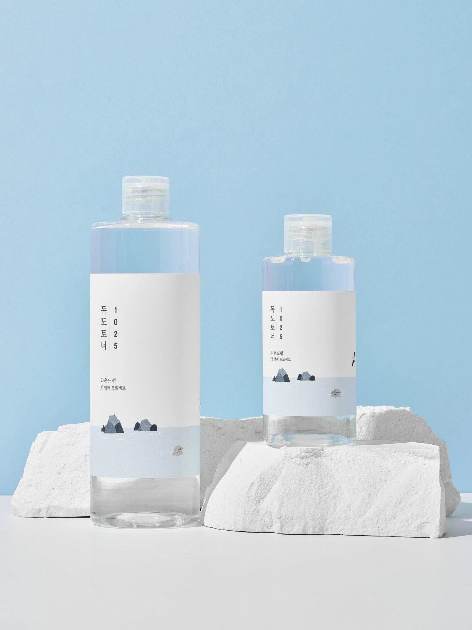 Two bottles of Round Lab 1025 Dokdo Toner with minimalist labels, arranged on white rocks against a soft blue background.