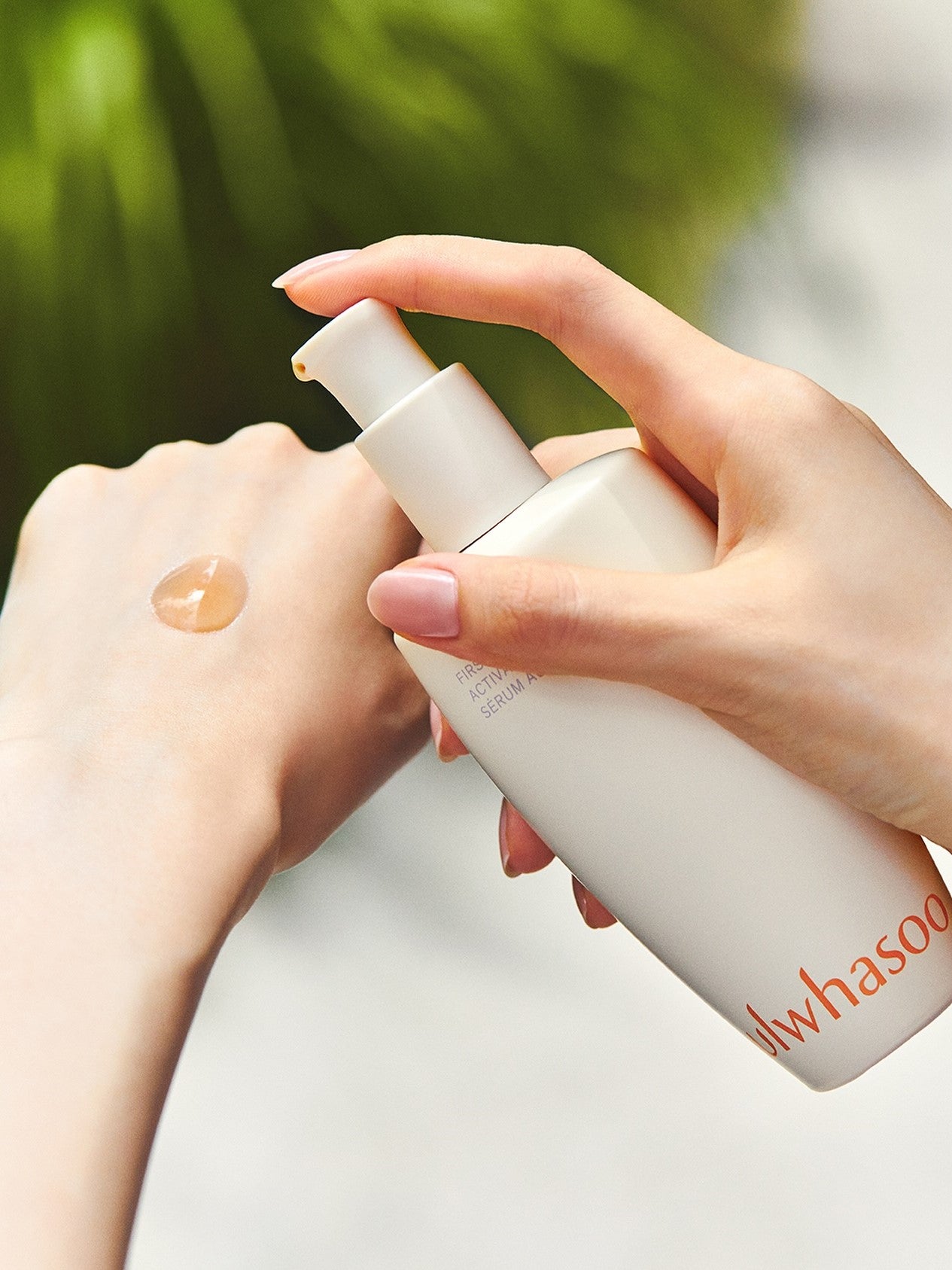 A hand dispenses a golden drop of Sulwhasoo First Care Activating Serum VI onto another hand, set against a blurred green background.