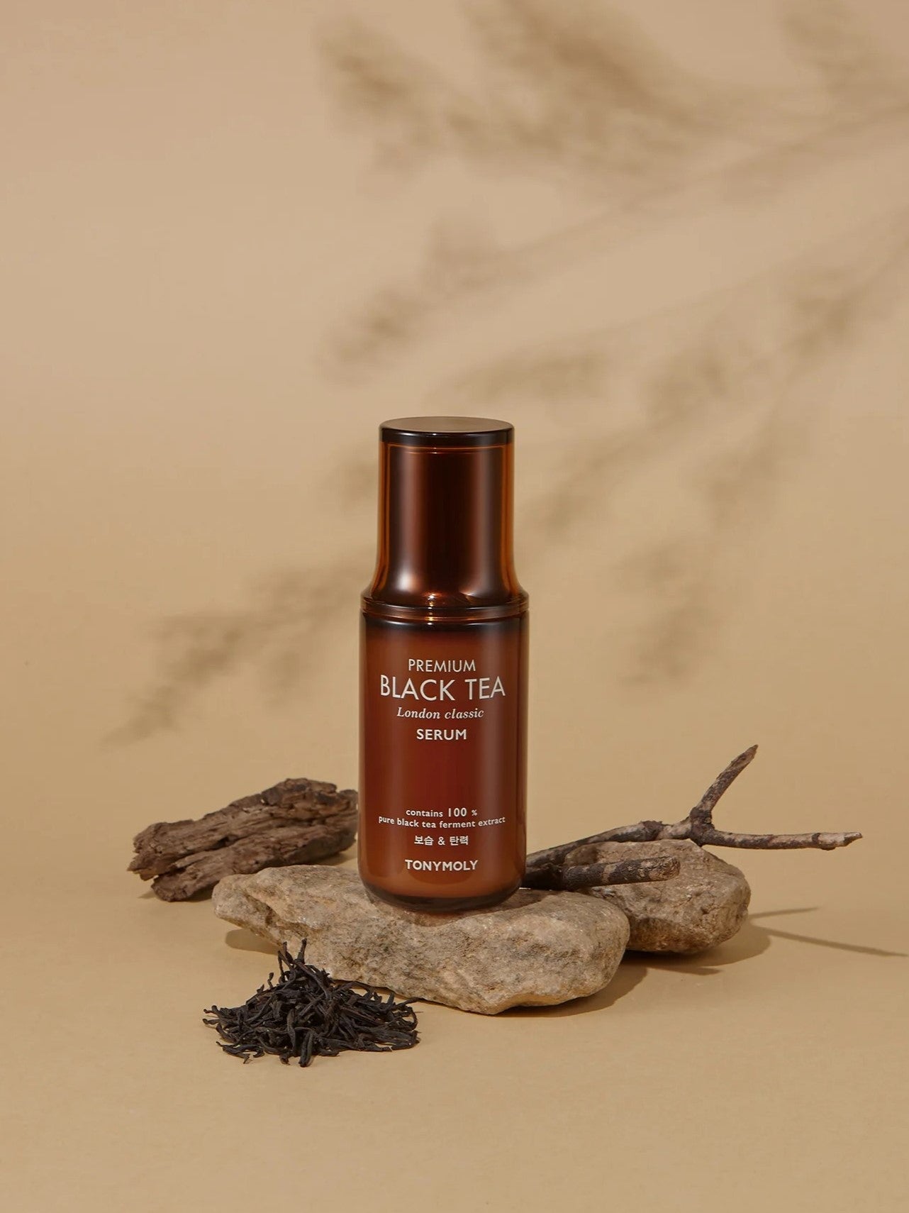 A bottle of Tonymoly The Black Tea London Classic Serum resting on stones, with loose tea leaves and twigs to highlight its natural ingredients, set against a beige backdrop.