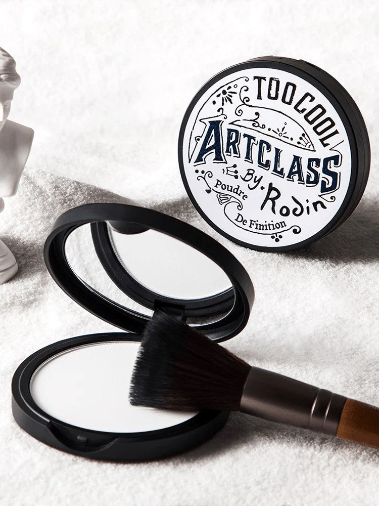 Close-up of Too Cool For School Art Class by Rodin Finish Setting Pact, paired with a makeup brush, resting on a soft white towel for a clean and polished aesthetic.