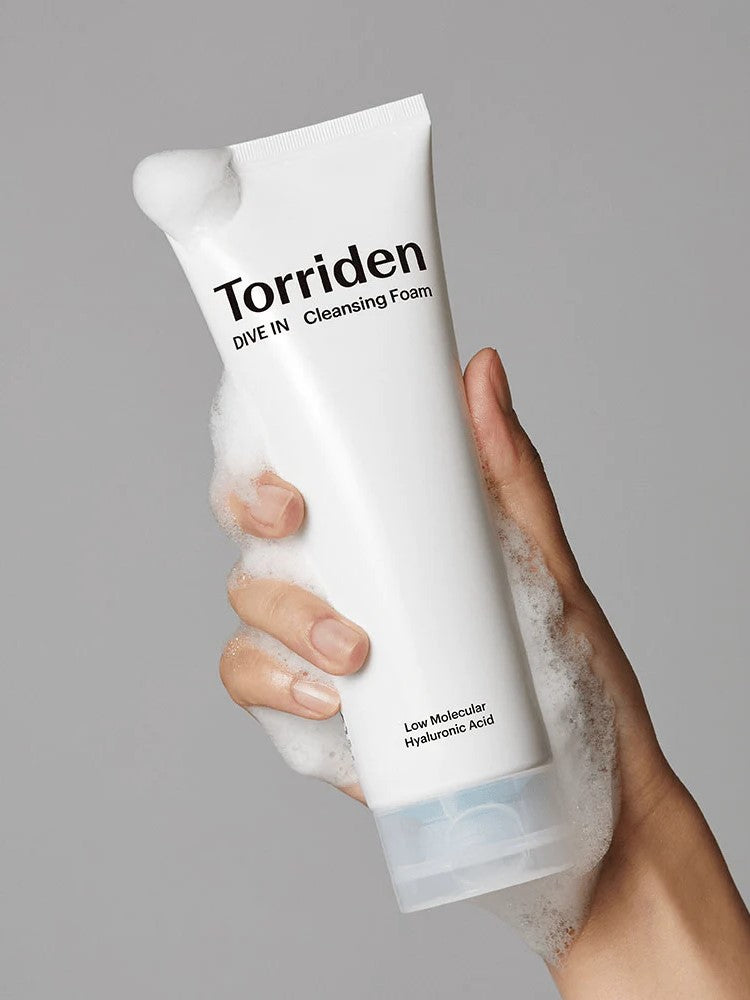 Hand holding a foamy tube of Torriden Dive In Cleansing Foam, with visible texture and labeled 'Low Molecular Hyaluronic Acid,' against a neutral background.