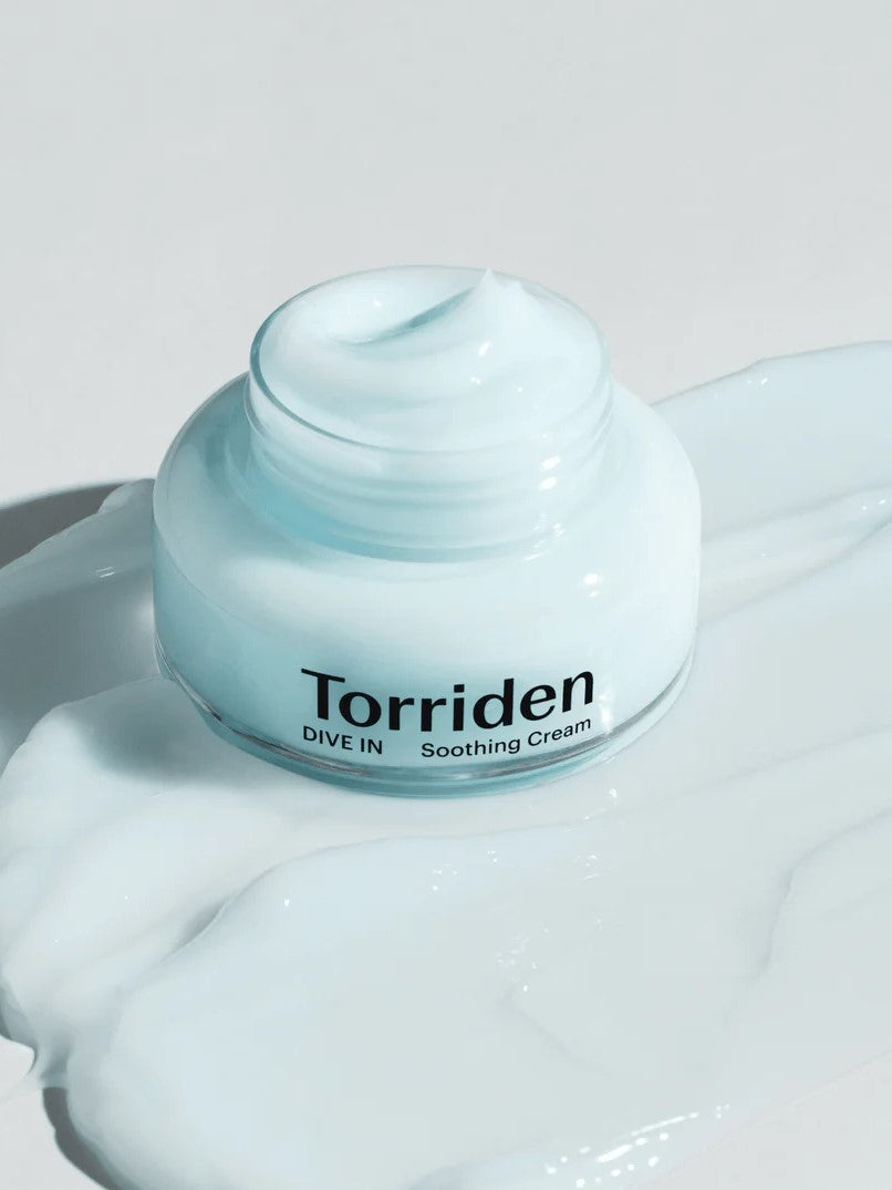 Jar of Torriden Dive In Low Molecular Hyaluronic Acid Soothing Cream on a reflective surface, surrounded by a creamy, light blue product spill.