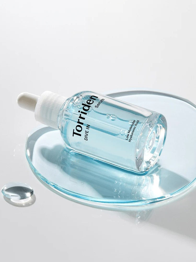 Clear blue serum bottle with a dropper, labeled 'Torriden Dive In,' resting on a glass plate surrounded by droplets of its lightweight texture.
