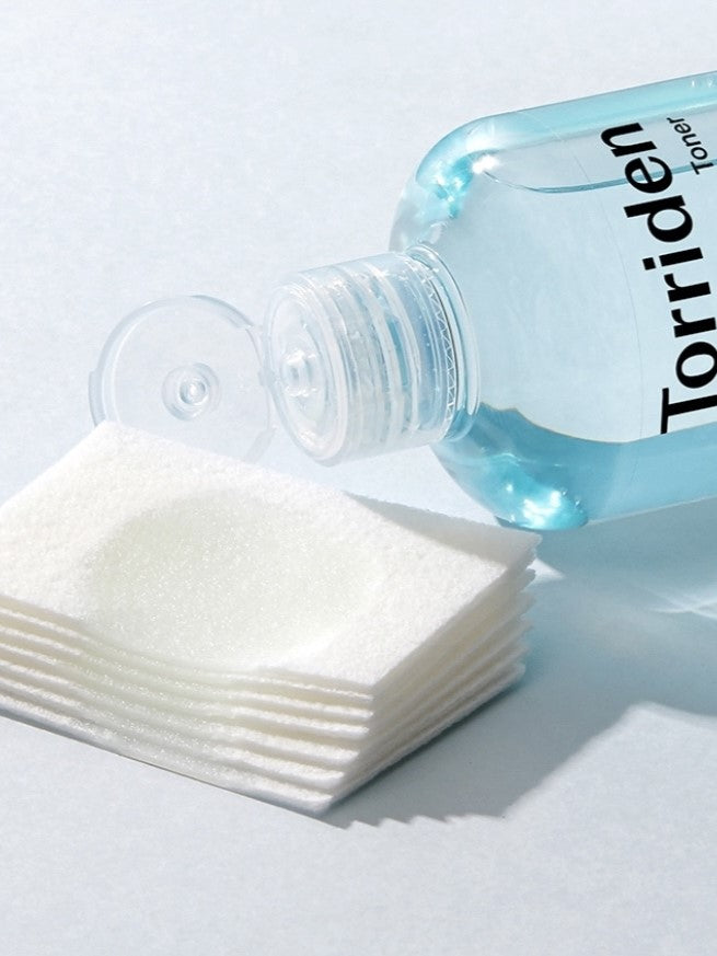 Bottle of Torriden toner next to stacked cotton pads, with a small amount of toner dispensed onto one pad.
