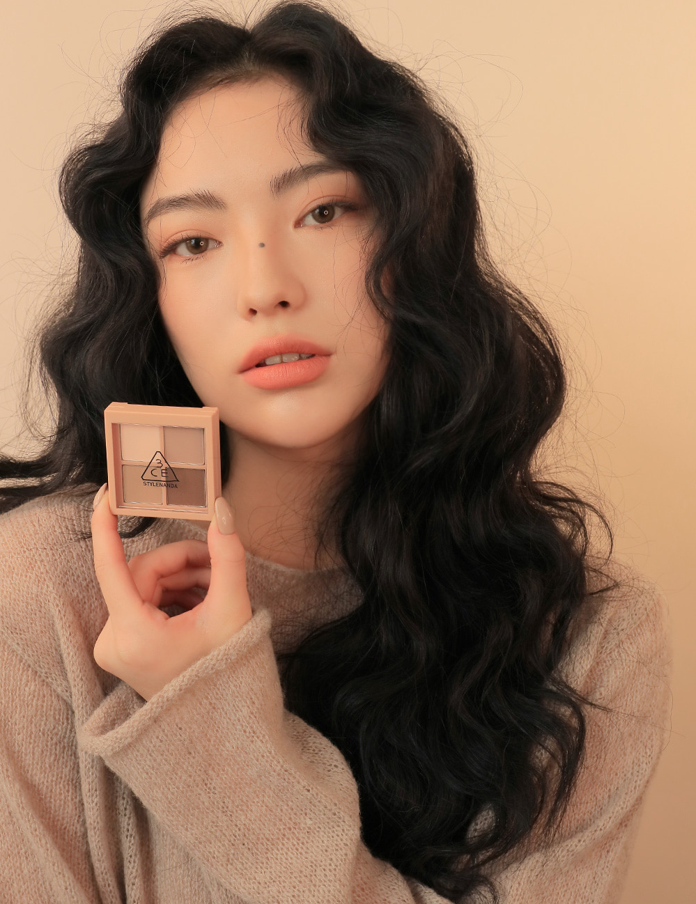 Model wearing a cozy beige sweater holding 3CE Mini Multi Eye Color Palette in Shade Flat Call, demonstrating its application and wearable look.