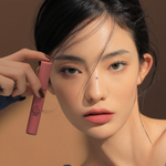 Model wearing a blue top against an earthy brown background featuring 3CE Velvet Lip Tint in Shade Cashmere Nude, emphasizing its soft texture and natural matte look.