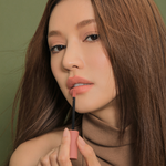 Model with long brown hair, sleeveless turtleneck, and multiple gold rings demonstrating 3CE Velvet Lip Tint in Shade Like Gentle, focusing on its smooth texture and rich color.
