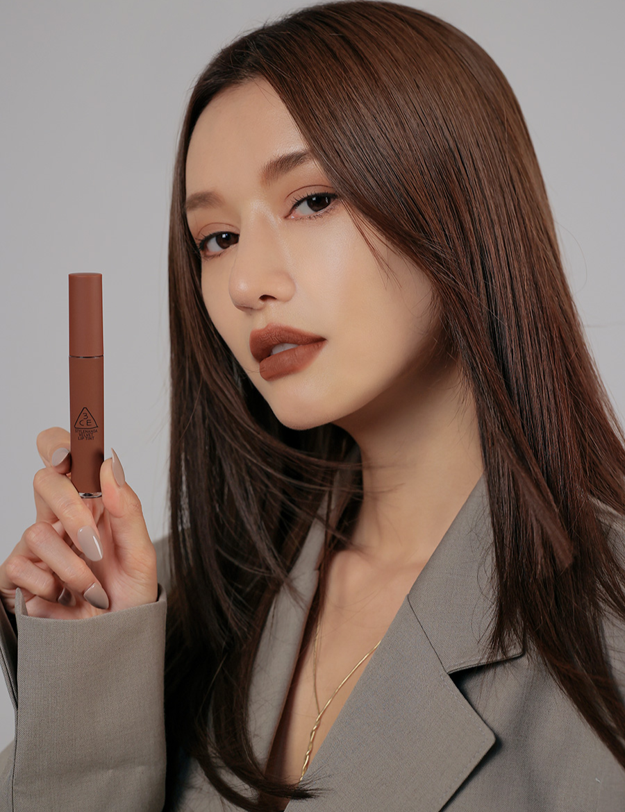 Model with long, sleek brown hair in a stylish gray blazer applying 3CE Velvet Lip Tint in Shade Bitten Hour, highlighting its velvety finish and intense color payoff.