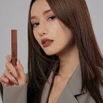 Model with long, sleek brown hair in a stylish gray blazer applying 3CE Velvet Lip Tint in Shade Bitten Hour, highlighting its velvety finish and intense color payoff.