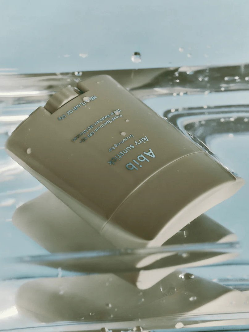 The Abib Airy Sunstick, a sleek beige sunscreen stick, floating on water with glistening droplets, highlighting its waterproof formula.