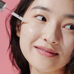 A model holds an Aestura Theracne 365 Active Serum dropper above her skin, with a soft pink background emphasizing the product’s light texture.