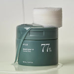 A sleek green jar labeled 'Anua Heartleaf 77% Clear Pad' with cotton pads resting on top, surrounded by a glossy liquid spill that highlights the essence of the pads.