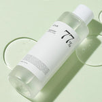 A clear bottle of Anua Heartleaf 77% Soothing Toner on a soft green background, with droplets of its lightweight texture visible on the surface.
