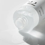 A partially tipped bottle of Anua Heartleaf 77% Soothing Toner spills a few drops onto a white surface, showcasing its lightweight texture.