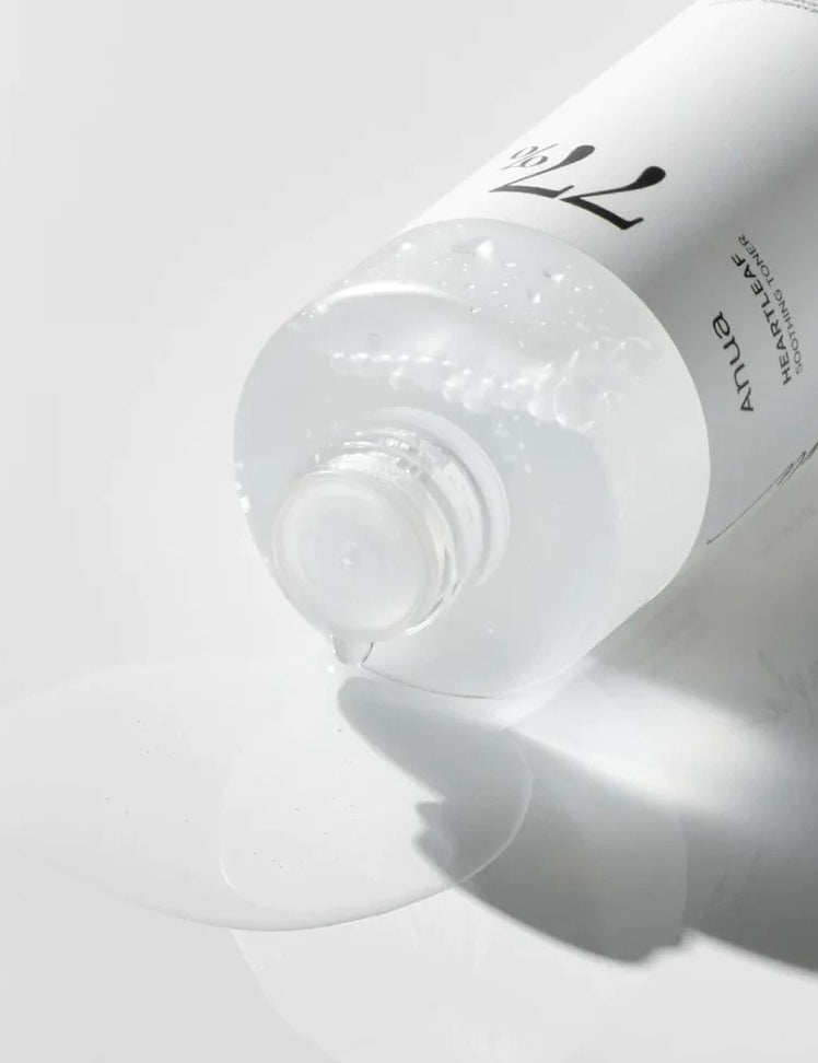 A partially tipped bottle of Anua Heartleaf 77% Soothing Toner spills a few drops onto a white surface, showcasing its lightweight texture.