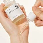 A hand holds a bottle of Anua Heartleaf Soothing Ampoule, with a dropper dispensing liquid onto the wrist.