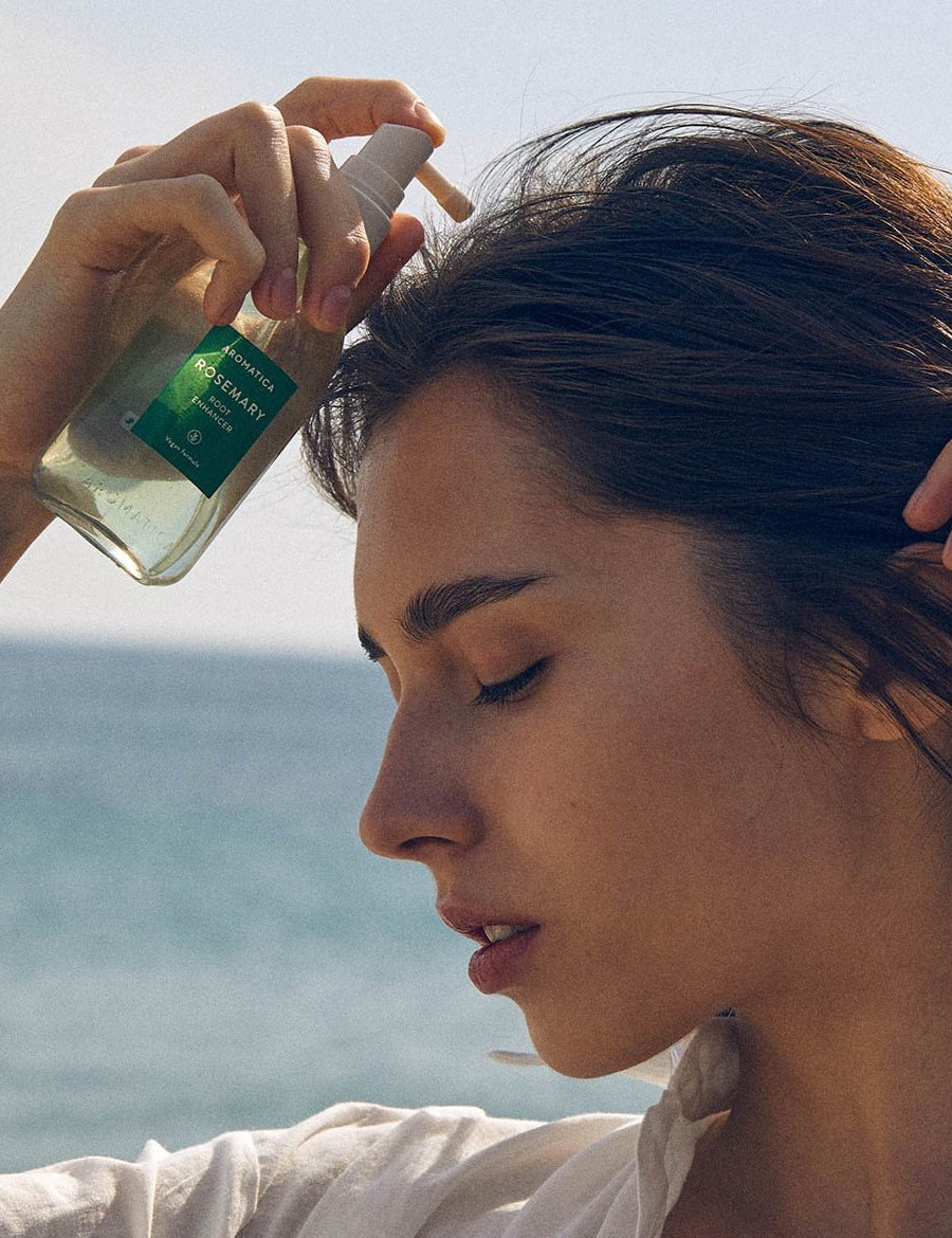 Model applying Aromatica Rosemary Root Enhancer scalp tonic, holding the clear pump bottle with ocean waves in the background.