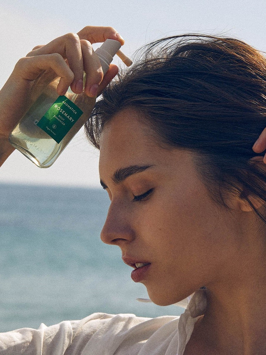 Model applying Aromatica Rosemary Root Enhancer scalp tonic, holding the clear pump bottle with ocean waves in the background.