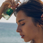 Model applying Aromatica Rosemary Root Enhancer scalp tonic, holding the clear pump bottle with ocean waves in the background.