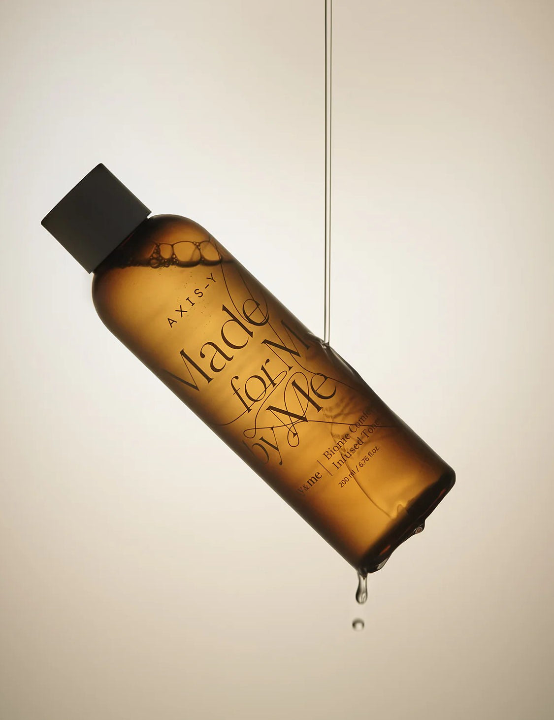 AXIS-Y Biome Comforting Infused Toner, suspended in air with a droplet falling from the bottom against a soft beige backdrop.