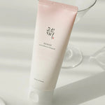 A tube of Apricot Blossom Peeling Gel from Beauty of Joseon, elegantly positioned on a reflective surface, with a glass in the background to enhance its aesthetic appeal.