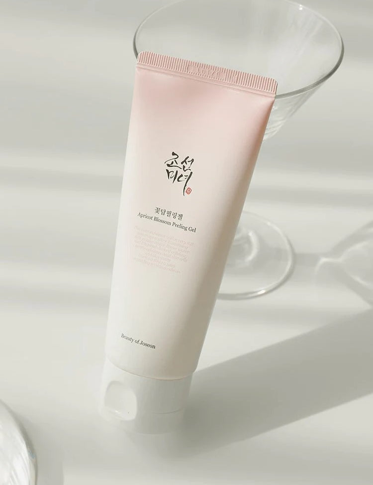 A tube of Apricot Blossom Peeling Gel from Beauty of Joseon, elegantly positioned on a reflective surface, with a glass in the background to enhance its aesthetic appeal.