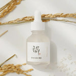 A glass dropper bottle of Beauty of Joseon Glow Deep Serum: Rice and Alpha Arbutin, surrounded by rice grains and soft, dried wheat on a light background, highlighting its key ingredients and nourishing properties.