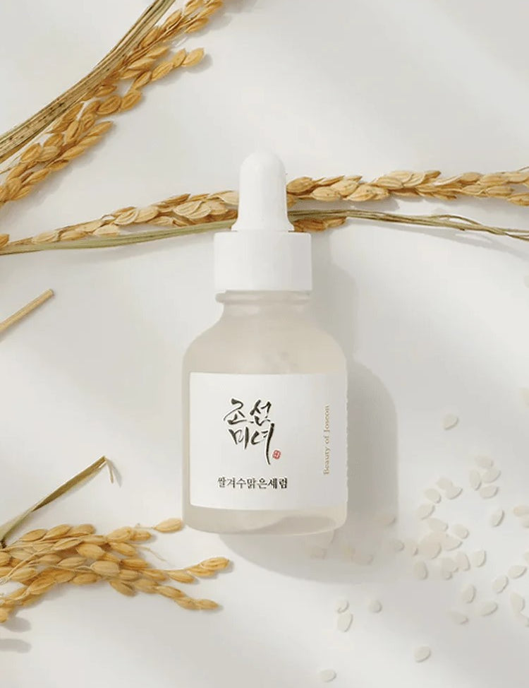 A glass dropper bottle of Beauty of Joseon Glow Deep Serum: Rice and Alpha Arbutin, surrounded by rice grains and soft, dried wheat on a light background, highlighting its key ingredients and nourishing properties.