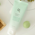 A mint-green tube of Beauty of Joseon Green Plum Refreshing Cleanser placed on a light surface, surrounded by fresh green plums and seeds, highlighting its natural ingredients.