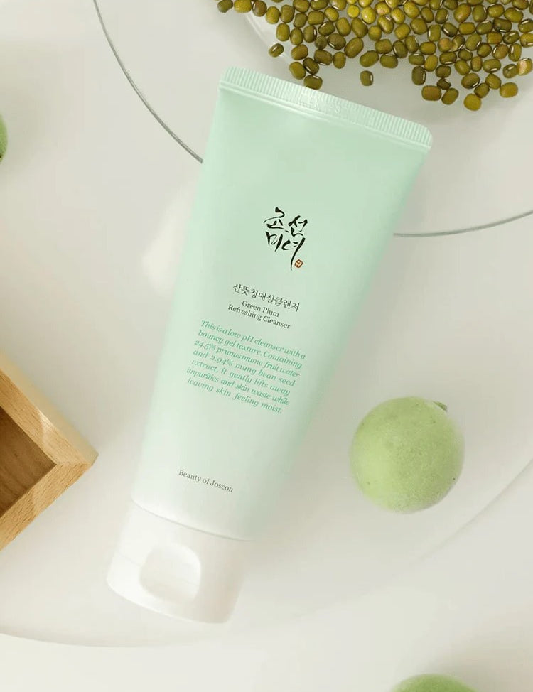 A mint-green tube of Beauty of Joseon Green Plum Refreshing Cleanser placed on a light surface, surrounded by fresh green plums and seeds, highlighting its natural ingredients.
