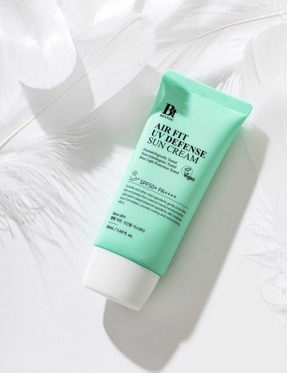 A mint green tube of Benton Air Fit UV Defense Sun Cream lying on a white background with feathers.